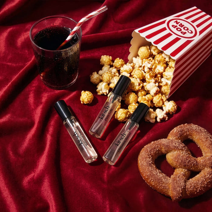 KITSCH Movie Theater Hair Perfume Trio Set — lifestyle flatlay on dark red velvet with movie theater food props: cola drink with straw, popcorn box, cinnamon pretzel alongside three kit·sch/ roller bottles — Buttered Popcorn, Fizzy Cola & Cinnamon Pretzel — Smell like a snack with three delectable scents — Wear each fragrance solo or layer them together to create your own signature scent