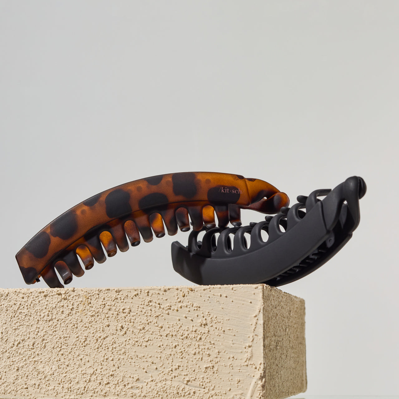 Two banana hair claw clips displayed on a textured beige block against a light gray background. One clip is tortoiseshell brown with black spots, and the other is solid black.