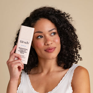 A model with curly dark hair holds a white tube of KitSch Moisturizing Leave-In Conditioner Spray next to her face against a solid beige background.
