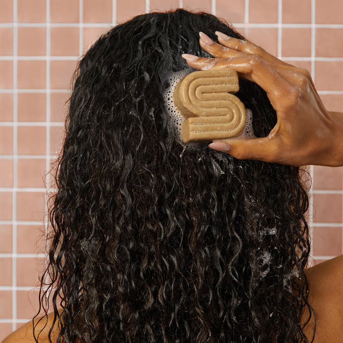 KITSCH Coconut Oil Solid Shampoo Bar — lifestyle back-view shot of female model with long curly hair applying shampoo bar to scalp in shower — pink tile background — Deeply cleanses and moisturizes kinky, curly, and coily hair from roots to ends — Formulated to support a healthier-feeling, more balanced scalp for stronger, revitalized hair — Perfect for Kinky, Curly, & Coily Hair