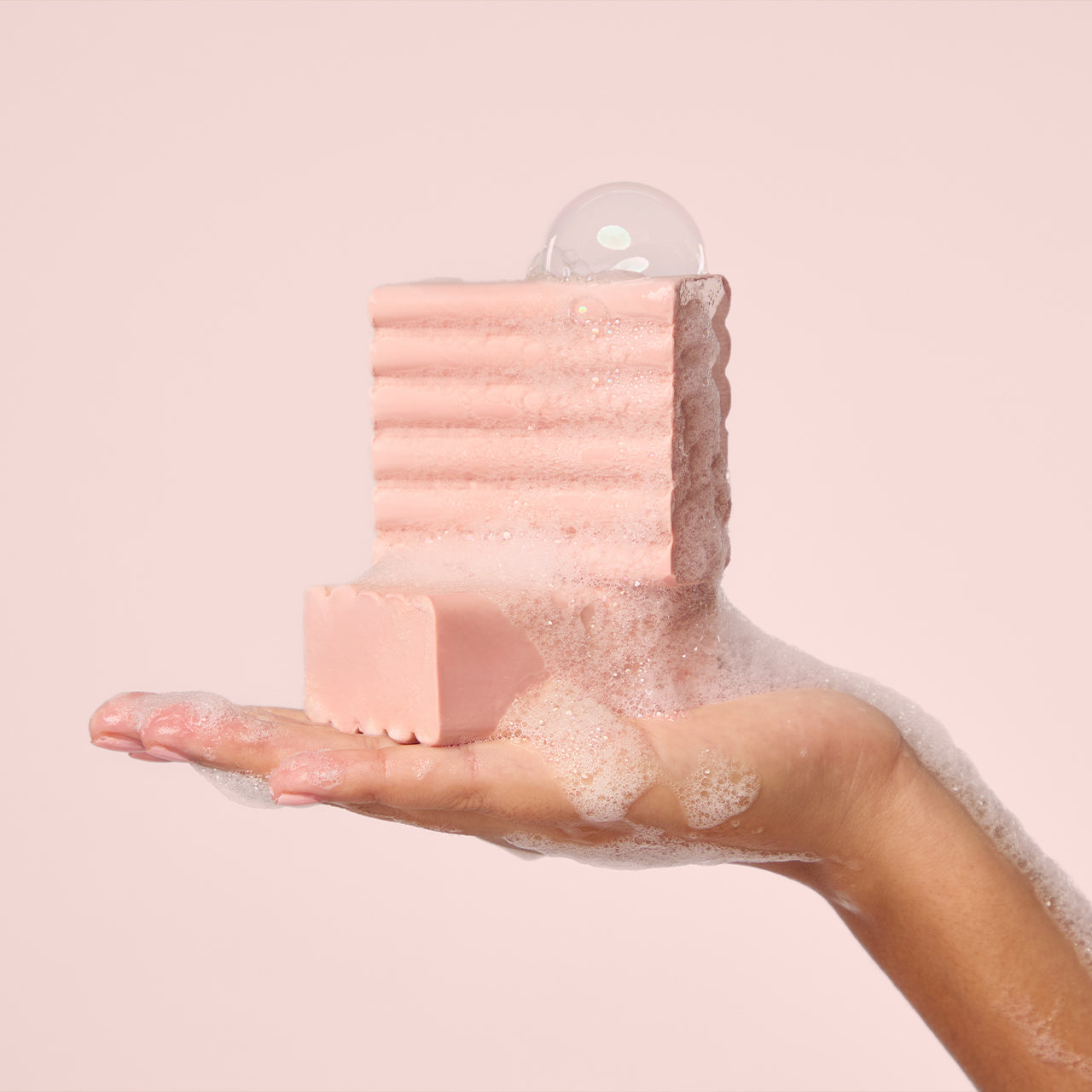 A hand holding a pink textured bath solid body wash with a matching bar resting in front, surrounded by white foam bubbles against a soft pink background.