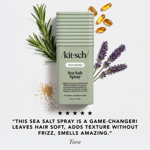 A bottle of kitsch Texturizing Sea Salt Spray centered on a light gray background, surrounded by rosemary sprigs, lavender stems, scattered sea salt grains, and amber capsules.
