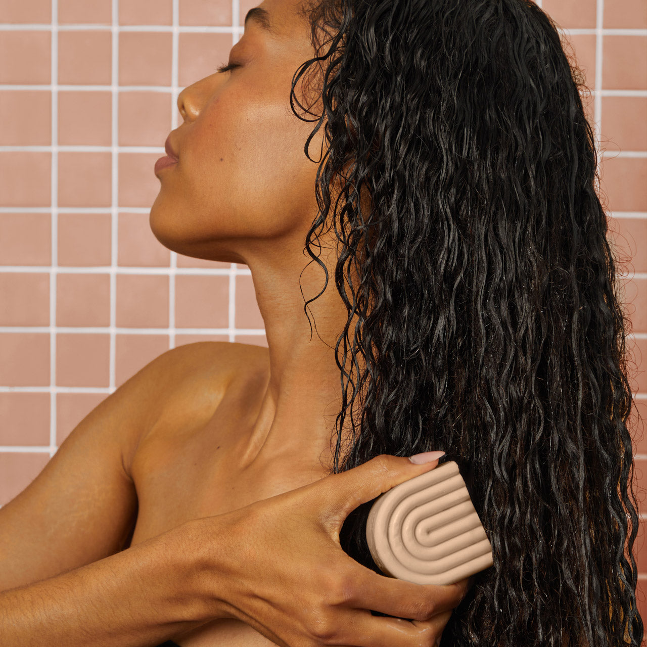 KITSCH Repairing Argan Oil Shampoo & Conditioner Bar Combo — lifestyle portrait of female model with long dark curly hair, side profile in pink tile bathroom, applying conditioner bar to hair — Enriched with PhytoVie® Defense to aid in recovery for damaged hair — Packed with antioxidant-rich argan oil to boost moisture without weighing it down — For damaged, weak, & brittle hair