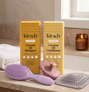 Kitsch Moisturizing Coconut Shampoo and Conditioner bottles on a bathroom counter, with a free soft violet wet/dry brush, a terracotta scalp exfoliator, and the rosewood net sponge as aa free welcome kit.