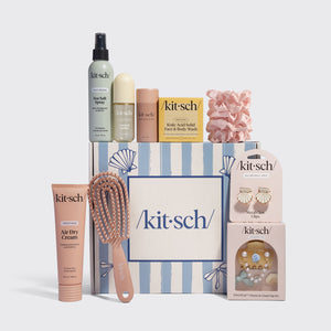 A Kitsch gift set arranged on a light gray background, featuring a striped gift box with seashell motifs, a pink hairbrush, a peach tube of Air Dry Cream, a pink scrunchie, a pink soap dish, a small packaged accessory set, and several bottles and boxes. Visible branding reads Kit-sch across multiple items.